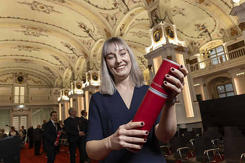 A woman smiles at the camera and holds up a red tube with his doctorate certificate