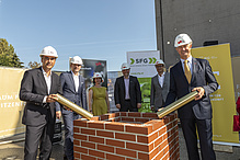5 men and a woman are standing on a building site - they are wearing white construction helmets. In the foreground are bricks.