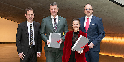 Vier Menschen stehen beieinander und schauen in die Kamera. Die beiden Personen in der Mitte halten Mappen in der Hand.