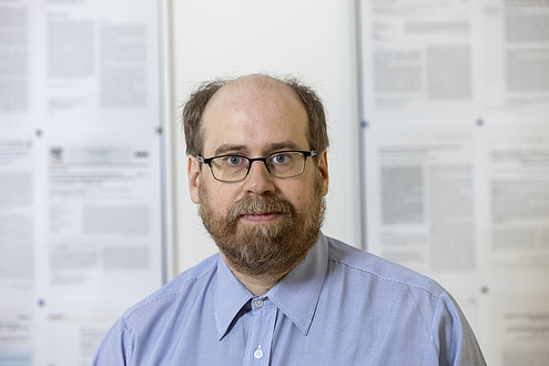 A man is standing in front of a wall with scientific essays pinned to it, looking into the camera.