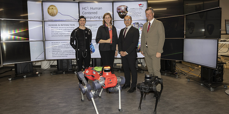 Four people stand behind two dog-like robots and look into the camera.