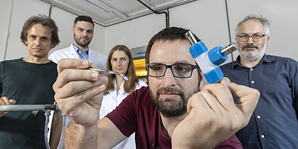 A man holds a pair of tweezers in his hand, four people stand in the background.