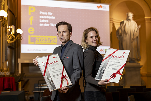 A man and a woman stand back to back and hold certificates declaring them as winners.