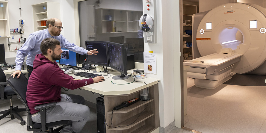 Two men are standing at a desk looking at a computer screen. In the background is an MRI machine.