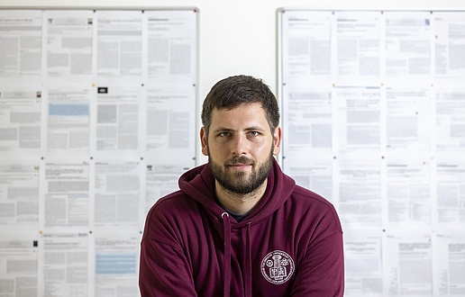 A man is standing in front of a wall with scientific essays pinned to it, looking into the camera.