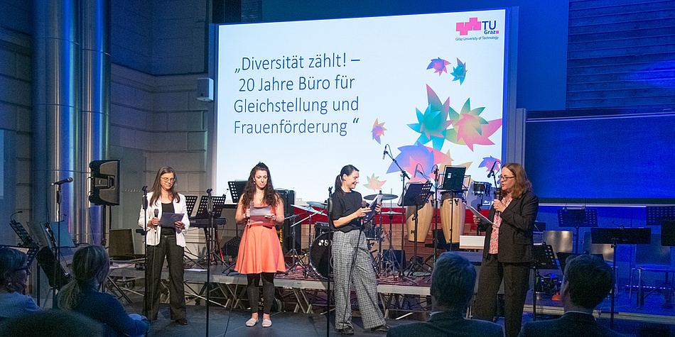 Vier Personen stehen vor einer Bühne, auf der Instrumente abgestellt sind. Sie halten Mikrofone und Zettel in den Händen. Im Hintergrund ist auf eine Leinwand "Diversität zählt! 20 Jahre Büro für Gleichstellung und Frauenförderung" projiziert.
