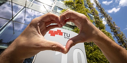 Mit Zeigefinger und Daumen wird ein Herz geformt - genau hinter dem Herz erscheint das Logo der TU Graz. 