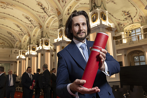 A man smiles at the camera and holds up a red tube with his doctorate certificate