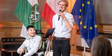 A young man in a wheelchair and a young man standing with a microphone in front of the flags of Styria, Austria and the EU.