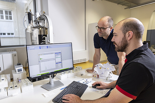 Two men sit in front of a computer and look at a diagram on the monitor.