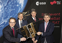 Four men in dark suits look into the camera and hold a satellite in their hands.
