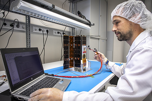 A man in a white coat is working on a laptop and a satellite in a laboratory.