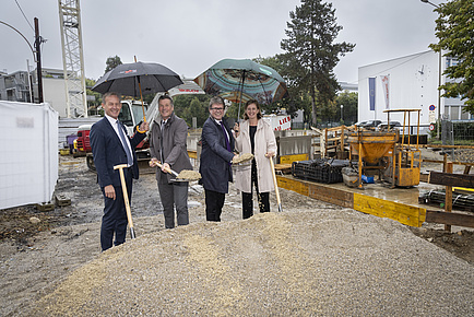 Four people, three men and one woman, stand under umbrellas in the rain. They are carrying shovels and smiling at the camera.