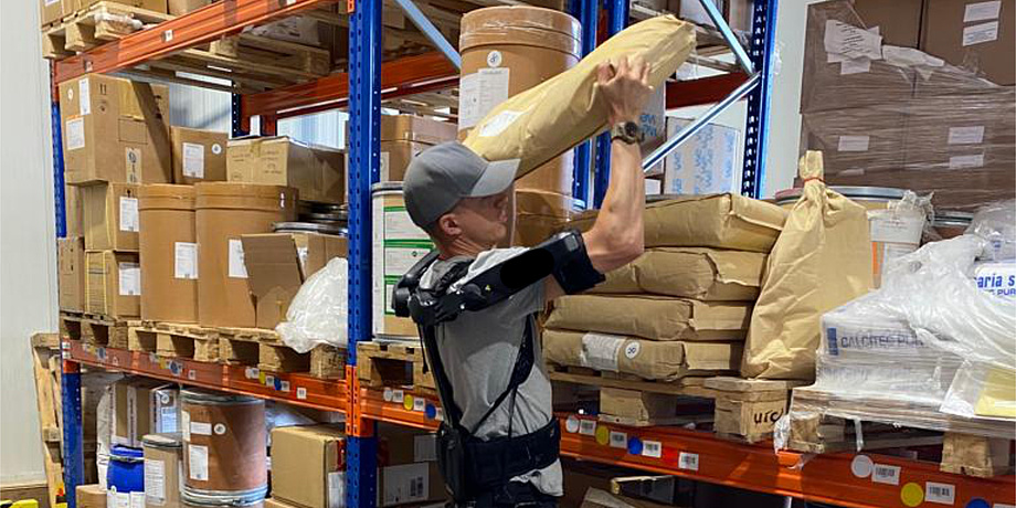 A man lifts a sack onto a shelf in a warehouse. He is wearing an exoskeleton over his clothes.