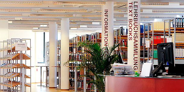 Bücherregale und ein Tresen mit Computer in einem großen Raum. An einer Säule steht Information, an einer anderen steht Lehrbuchsammlung.