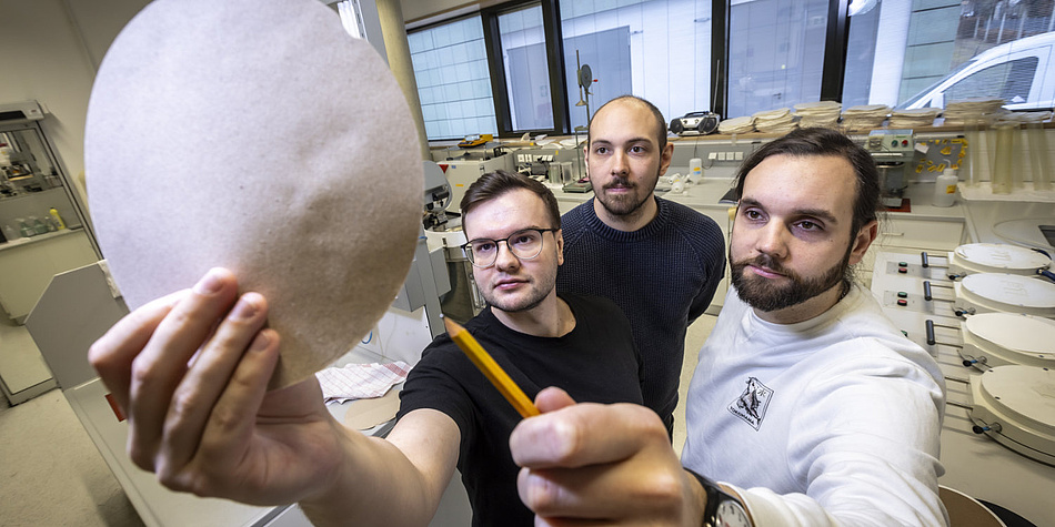 Three men are standing together looking at a round piece of paper that one of them is holding up.