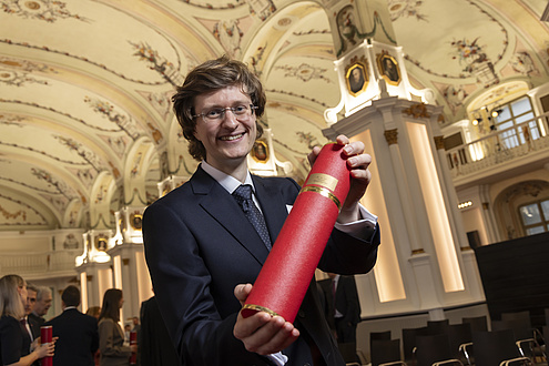 A man smiles at the camera and holds up a red tube with his doctorate certificate