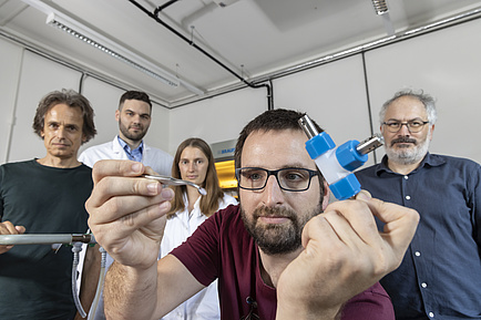 A man holds a pair of tweezers in his hand, four people stand in the background.