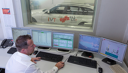 Ein Herr im weißen Hemd beobachtet fünf Bildschirme und im Hintergrund durch ein Fenster ein weißes Auto