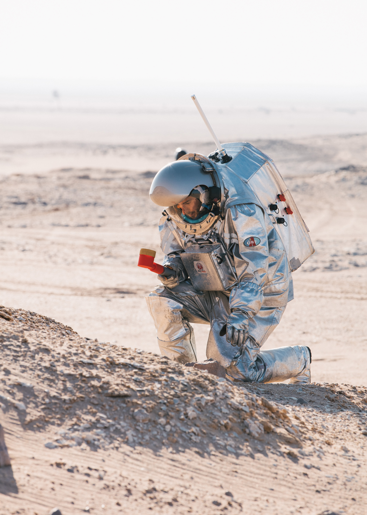 A man in a space suit kneeling takes samples in the red desert sand. He holds a tool in one hand.